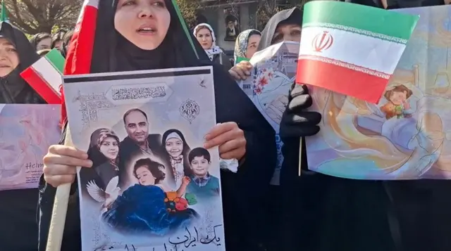 Mulheres no funeral da família de Helma seguram bandeiras do Irã e cartazes com fotos da família