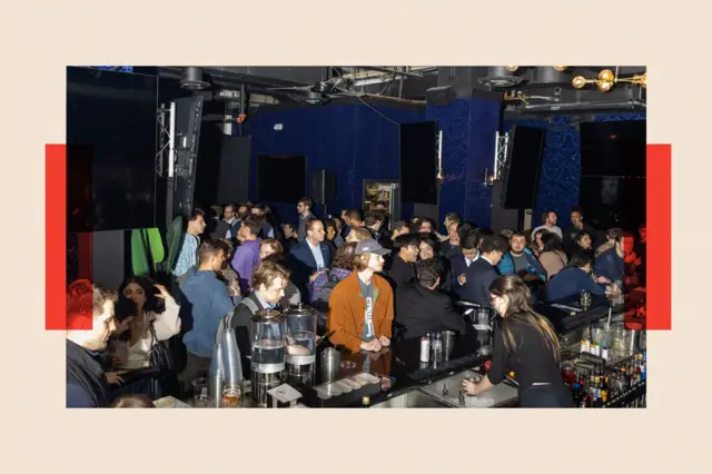 A queue for the bar in a dark room. Most of the customers are young men, 