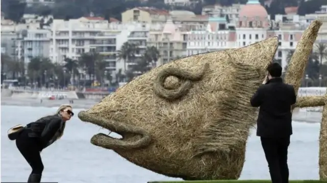 Obinrin kan n wo ẹja ti wọn fi igi gbẹ́, ọkunrin kan naa duro lẹyin, o n wo ẹja yii.