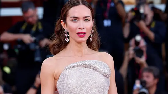 Emily Blunt está de pie sobre una alfombra roja. Lleva un vestido plateado y pendientes.