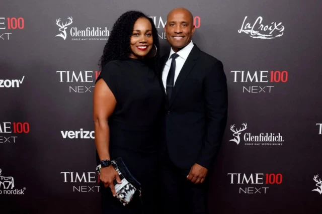 Мужчина в костюме, женщина в черном платье. На стене за ними рекламные надписи