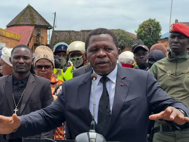 Un homme vêtu d’un costume bleu, chemise blanche et cravatte noire, s’adresse à la foule. Derrière lui, des hommes en tenues civiles et militaires.