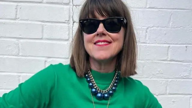Caroline wears a bright green jumper, two gold necklaces and a large metallic blue beaded necklace. She is also wearing sunglasses and bright red lipstick. Her hair is brown and shoulder-length with a fringe.