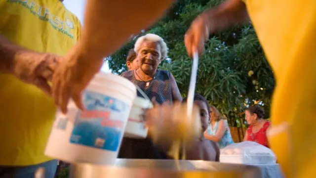 Personas con camisetas amarillas distribuyen comida en Ciudad de Natal, Brasil. En el fondo se ve una anciana.
