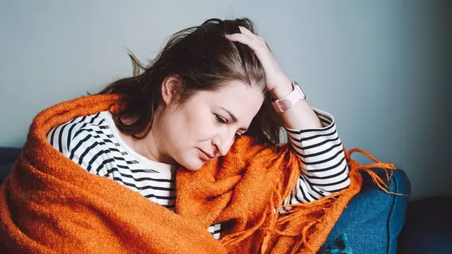 Una mujer con una camiseta blanca con rayas negras envuelta en una manta naranja está sentada cabizbaja en un sofá azul