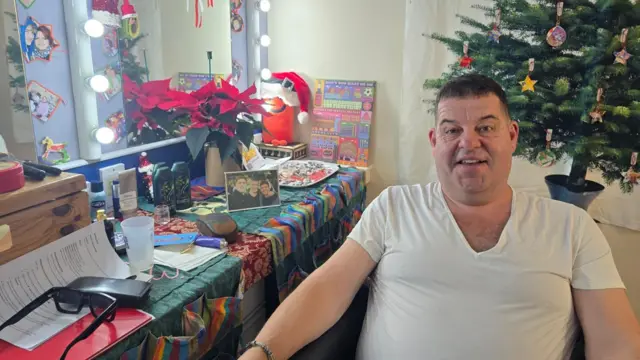 Damian Williams estaba sentado en su camerino con una camiseta blanca con un árbol de Navidad de fondo y sus gafas, baño de burbujas, fotos enmarcadas y un espejo iluminado a su lado.