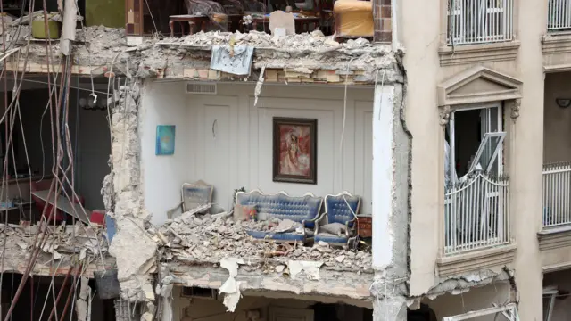 Bloco de apartamentos residenciais parcialmente destruído em Teerã, sem a parede externa e com um sofá azul em um cômodo repleto de escombros e um quadro ainda pendurado na parede