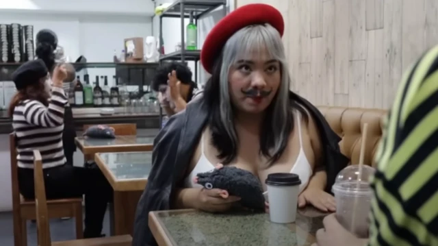 Женщина с нарисованными усами в красном берете сидит в кафе, на заднем плане два мима