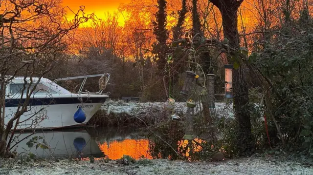 Un canal o río con un barco amarrado. El cielo es naranja, lo que se refleja en el agua. Hay escarcha en las orillas.