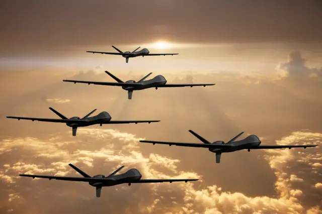 Cinq drones militaires, semblables aux MQ-9 Reaper de surveillance et de combat, volent en formation décalée au-dessus d'un coucher de soleil flamboyant.