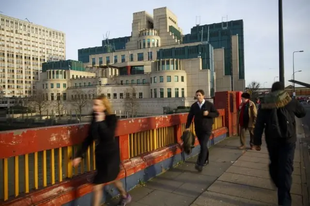 MI6 genel merkezinin nehir üzerindeki köprüden çekilmiş fotoğrafı. Kadrajda yürüyen insanlar var.