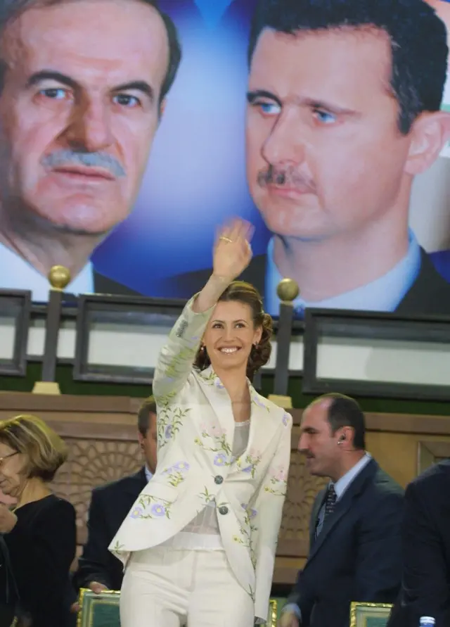 Asma al Assad saluda sonriente, detrás de ella hay una foto de Bashar al Assad y su padre Hafez al Asad