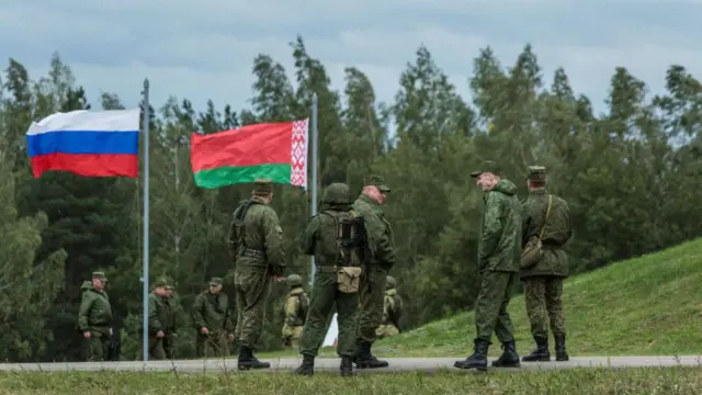 На фоне леса стоят военные в камуфляже. Рядом развеваются флаги России и Беларуси