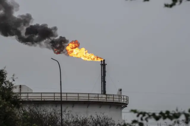 Зроблене здалеку фото нафтового заводу у Венесуелі. Видно колючий дріт на паркані, резервуар, а за ним труба, над якою горить вогонь.