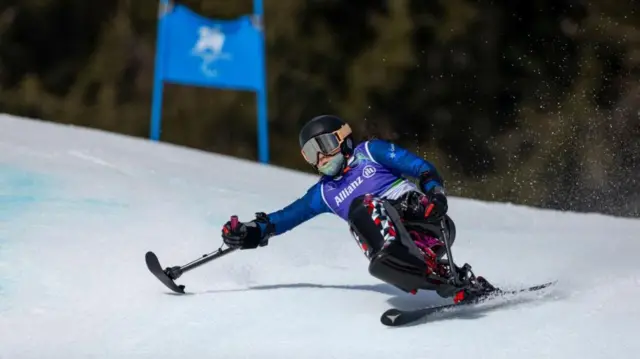 ヘルメットをかぶりゴーグルを着け、ゼッケンを着けた村岡が、ストック代わりに端がスキーのようになっている用具アウトリガーを使いながら滑っている