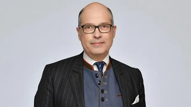 Un hombre con gafas, chaqueta, chaleco, corbata y pañuelo blanco en el bolsillo del pecho de la chaqueta. 