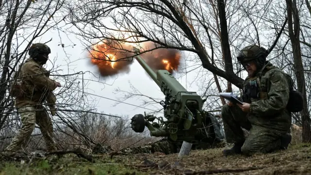 Dwóch ukraińskich żołnierzy kryje się, gdy z małego lasu armata artyleryjska ostrzeliwuje pozycje rosyjskie. Z lufy armaty wydobywa się ogień i dym.