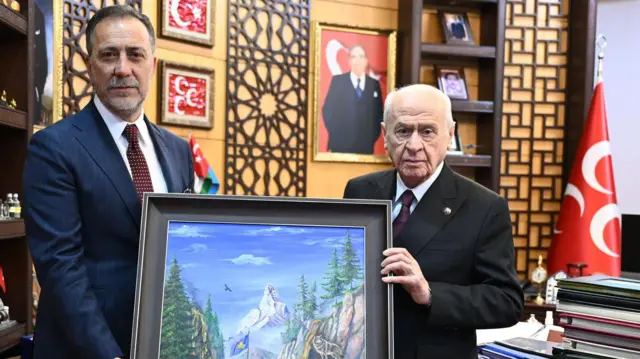 Volkan YIlmaz ile Devlet Bahçeli ayakta ellerinde bir tablo tutarak kameraya bakıyor