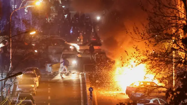 Un incendie fait rage là où des voitures sont garées à Téhéran, le 8 janvier 2026. Des gens sont rassemblés à proximité. Un arbre se trouve au premier plan.