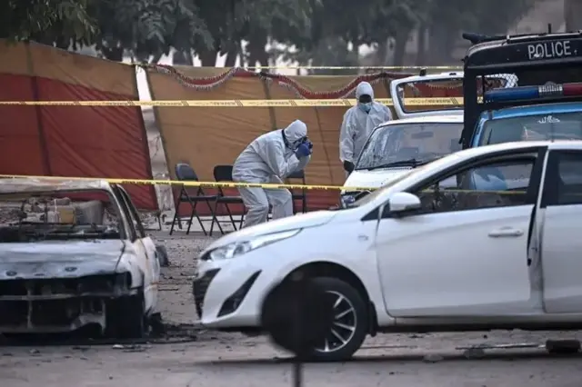 බෝම්බය පුපුරා ගිය ස්ථානය නිරීක්ෂණය කරන පිරිසක් ඡායාරූපයේ දැක්වේ