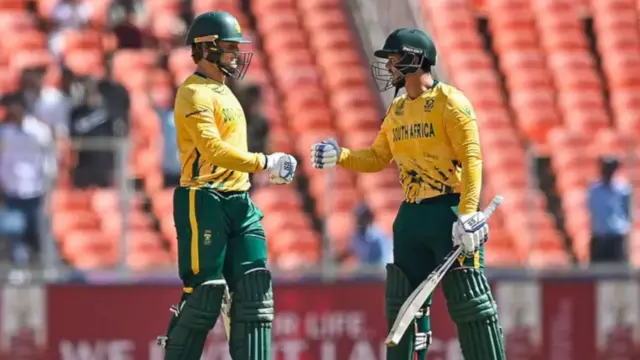 دو بازیکن افریقای جنوبی هنگام توپ‌زنی در بازی با افغانستان. آنها پیراهن‌های زرد رنگ و شلوار و کلاه سبز رنگ به تن دارند. 