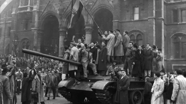 Na czarno-białej fotografii grupa protestujących trzyma flagę na szczycie czołgu przed budynkiem parlamentu w Budapeszcie podczas powstania węgierskiego przeciwko rządom sowieckim w październiku 1956 r. 