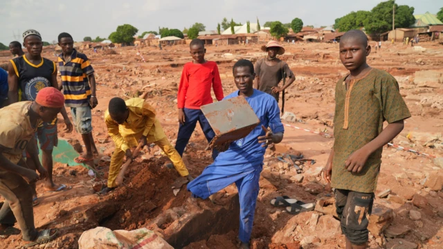Group of boy swey bin dey dig di remains, some look straight, one hold laptop