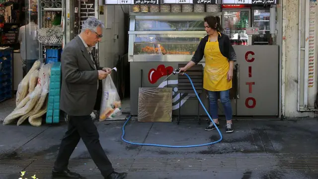 Мужчина проходит мимо, женщина моет тротуар из шланга возле небольшого ресторана в Тегеране.