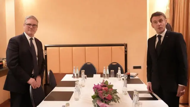 Лидеры Британии и Франции Ким Стармер и Эммануэль Макрон стоят по обе стороны большого стола и смотрят в объектив