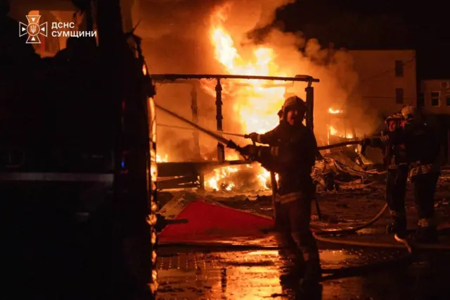 Рятувальники ДСНС ліквідовують масштабну пожежу в нічний час. На задньому плані видно потужне полум’я, яке охопило будівлю або промисловий об’єкт, та густий дим. Пожежники працюють зі шлангами біля пожежного автомобіля, а мокре покриття відбиває яскраве світло вогню.