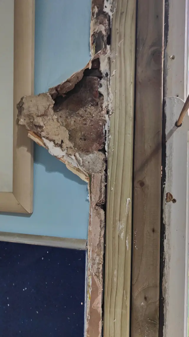 Dos listones de madera se interponen entre la ventana y la pared. La pared está dañada y algunos trozos se están desprendiendo.