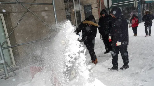 عمال يرتدون سترة سوداء وسروالاً أسود، وقفازات ويغطون رؤوسهم بعظاء رأس واقٍ من البرد، بينما يستخدمون آلات لإزالة الثلج من رصيف المشاة بينما تمتد العاصفة الشتوية 