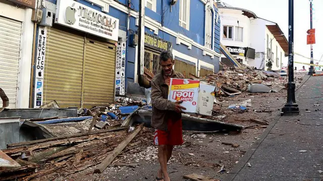 Un hombre camina con una caja de cartón en las manos por una calle donde hay escombros por todos lados.