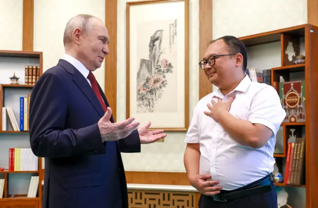 Владимир Путин говорит с китайским инженером (невысоким мужчиной в очках и белой рубашке с коротким рукавом) на фоне книг и картины.