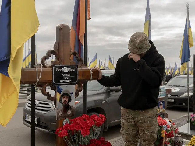 Чоловік у камуфляжних штанах і чорному худі стоїть біля дерев’яного хреста, схиливши голову й торкаючись рукою до нього. Перед хрестом стоять червоні гвоздики. Навколо встановлені численні українські прапори, а на тлі видно припарковані автомобілі та похмуре небо.