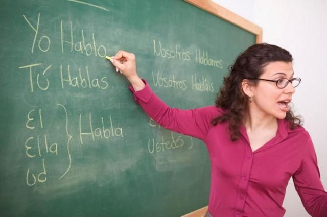 Professora branca de cabelo preto encaracolado nos ombros escreve palavras em espanhol em uma lousa enquanto fala