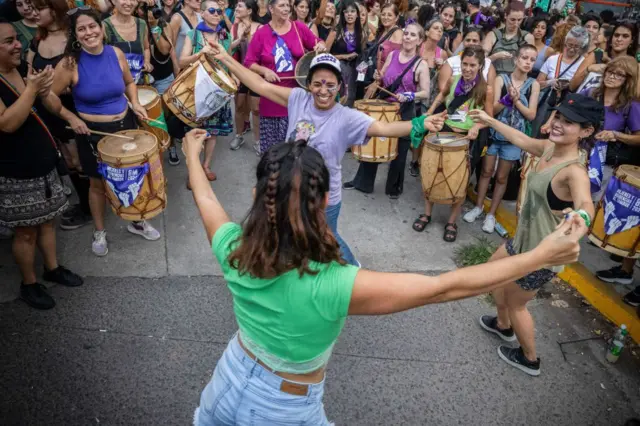Tres mujeres bailan tomándose de las manos en lo que parece ser una manifestación feminista. En el fondo, otras mujeres las miran y tocan tambores.