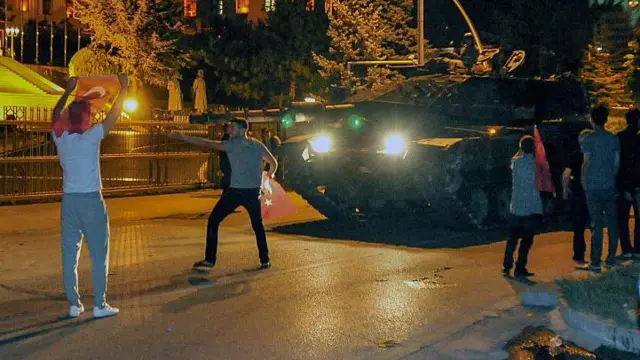 15 Temmuz 2016 gecesinden İstanbul'da çekilmiş bir fotoğraf. Bir tank yolda ilerliyor. Önünde ise darbe girişimine karşı çıkan iki kişi ellerinde Türk bayraklarıyla duruyor. Tankın yanında da küçük bir kalabalık görülüyor. 