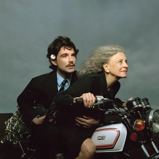Um homem jovem (Jão) e uma mulher idosa (Marisa Sabino dos Santos) estão sobre uma motocicleta branca, ambos vestidos de preto e olhando para a frente. O homem usa terno, camisa azul, gravata e uma flor branca atrás da orelha, enquanto a mulher, de cabelos grisalhos, segura o guidão da moto. Na parte traseira do veículo há um buquê de flores brancas, e o fundo é formado por um céu cinzento.