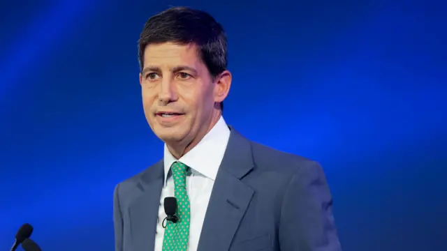 Кевин Уорш, мужчина средних лет с темными короткими волосами и карими глазами, без бороды, в пиджаке с зеленым галстуком.