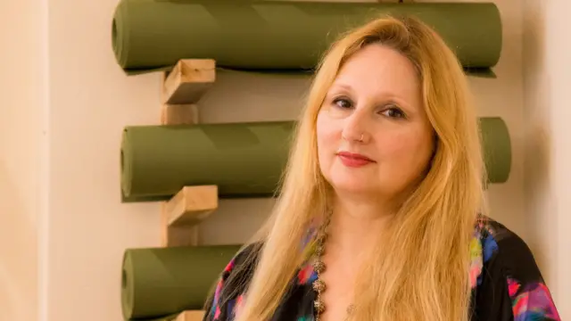 Florence Achery tem cabelos longos e loiros. Atrás dela há vários tapetes de yoga pendurados na parede 