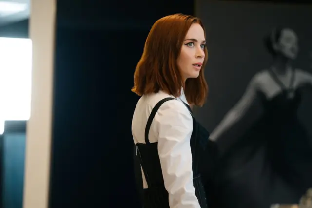 Side-on image of Emily Blunt wearing a white shirt and black dress.