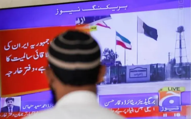 A man looks at a TV screen showing Pakistani and Iranian flags flying