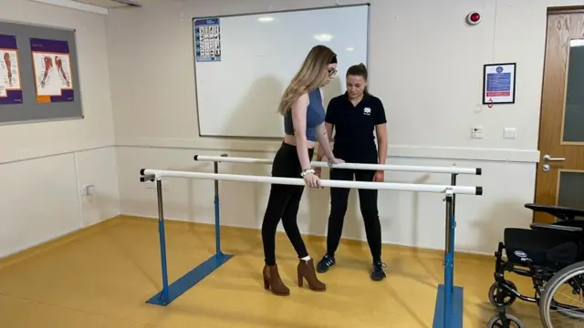 Megan está em pé segurando em duas barras. Uma mulher está ao lado dela e há uma cadeira de rodas na sala. 