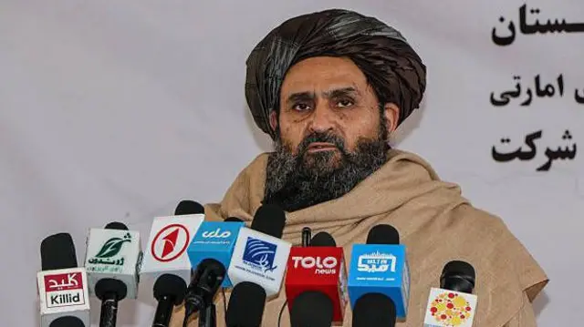 تصویر ملا عبدالغنی برادر. او ریش سیاه دارند و دستار سیاه بر سر کرده است. به دورش پتوی زرد پیچیده و در پشت مایک برخی رسانههای محلی افغانستان ایستاده است.