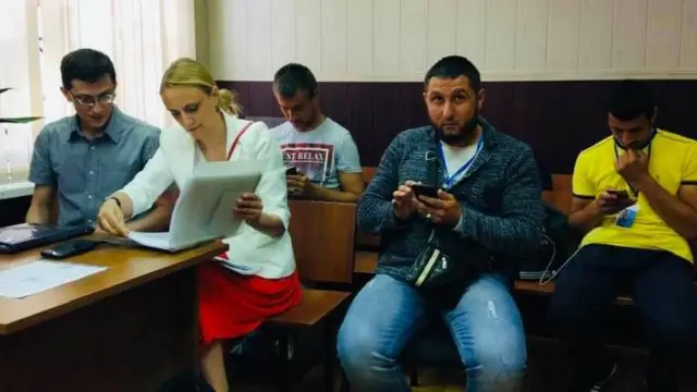 женщина с папкой документов и четверо мужчин сидят в зале суда