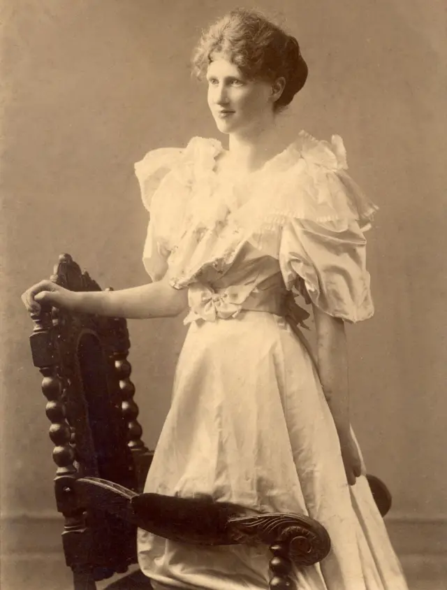 
Eglantyne com um vestido longo e claro, apoiada em uma cadeira.
