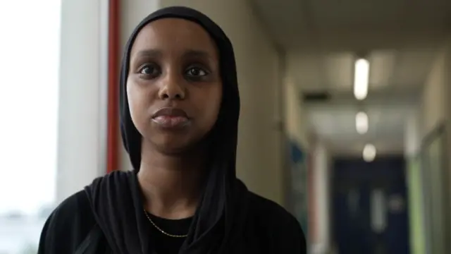 Saynab Axmed oo 17 jir ah waxay taagan tahay meel daaqad u dhow. Waxay eegaysaa kamaradda. Waxay si xarrago leh ugu lebbisan tahay shalmad madow, Cabbaayad madow iyo katiinad dahab ah.