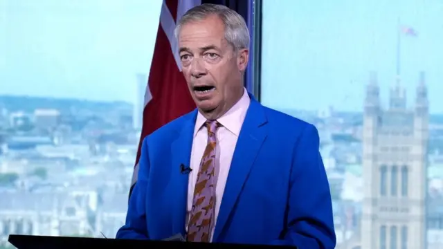 イギリス議会議事堂などの建物が見える窓を背に、青いジャケットに淡いピンクのシャツ、茶色い魚の絵がデザインされたピンク色のネクタイを着けたファラージ氏が前を見て話している