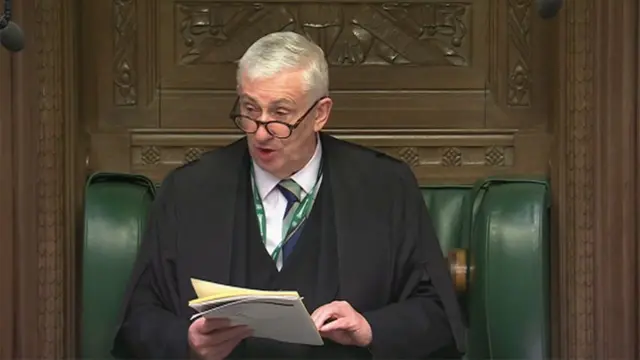 彫刻の施された茶色い木のパネルと緑色の皮の背もたれがはめこまれている議長席に座り、ワイシャツとネクタイの上に黒いガウンを着た白髪の議長が、眼鏡をかけ、手元のメモを読みながら発言している